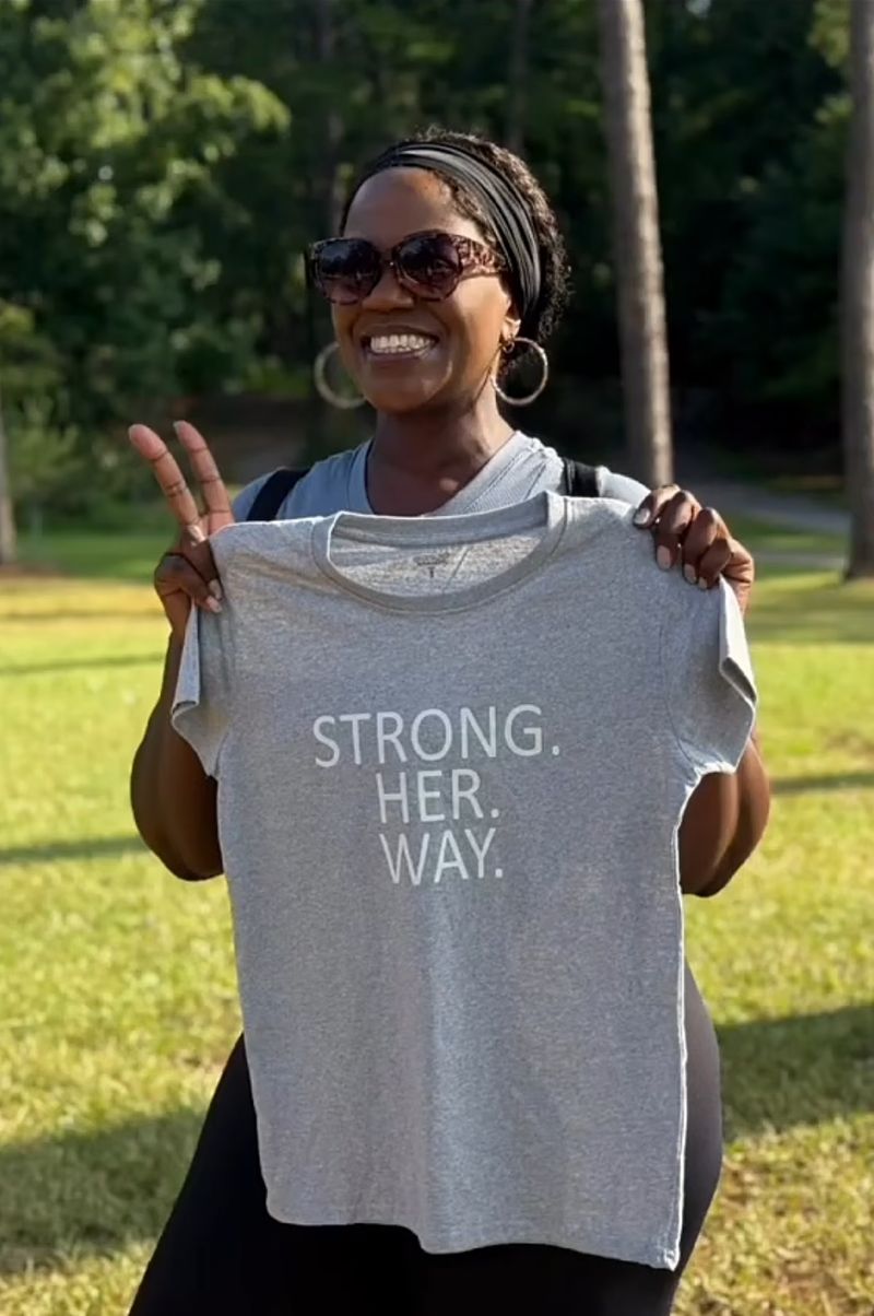 A StrongHERway member smiling and holding up a Strong. Her. Way. tee in the park